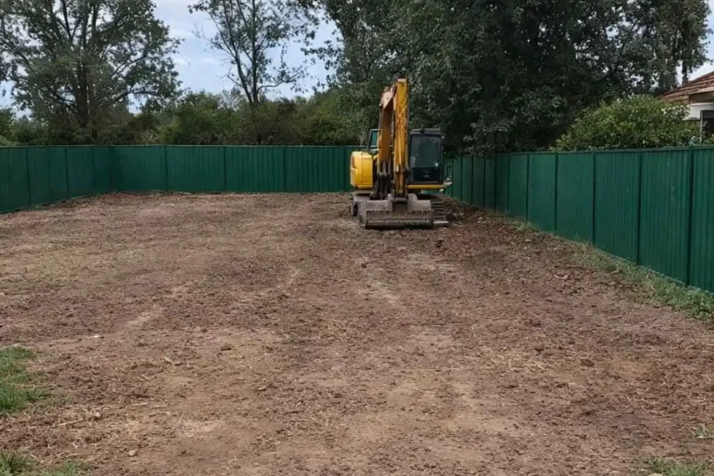 Excavator levelling cleared residential demolition site in Sydney with green Colorbond fencing.